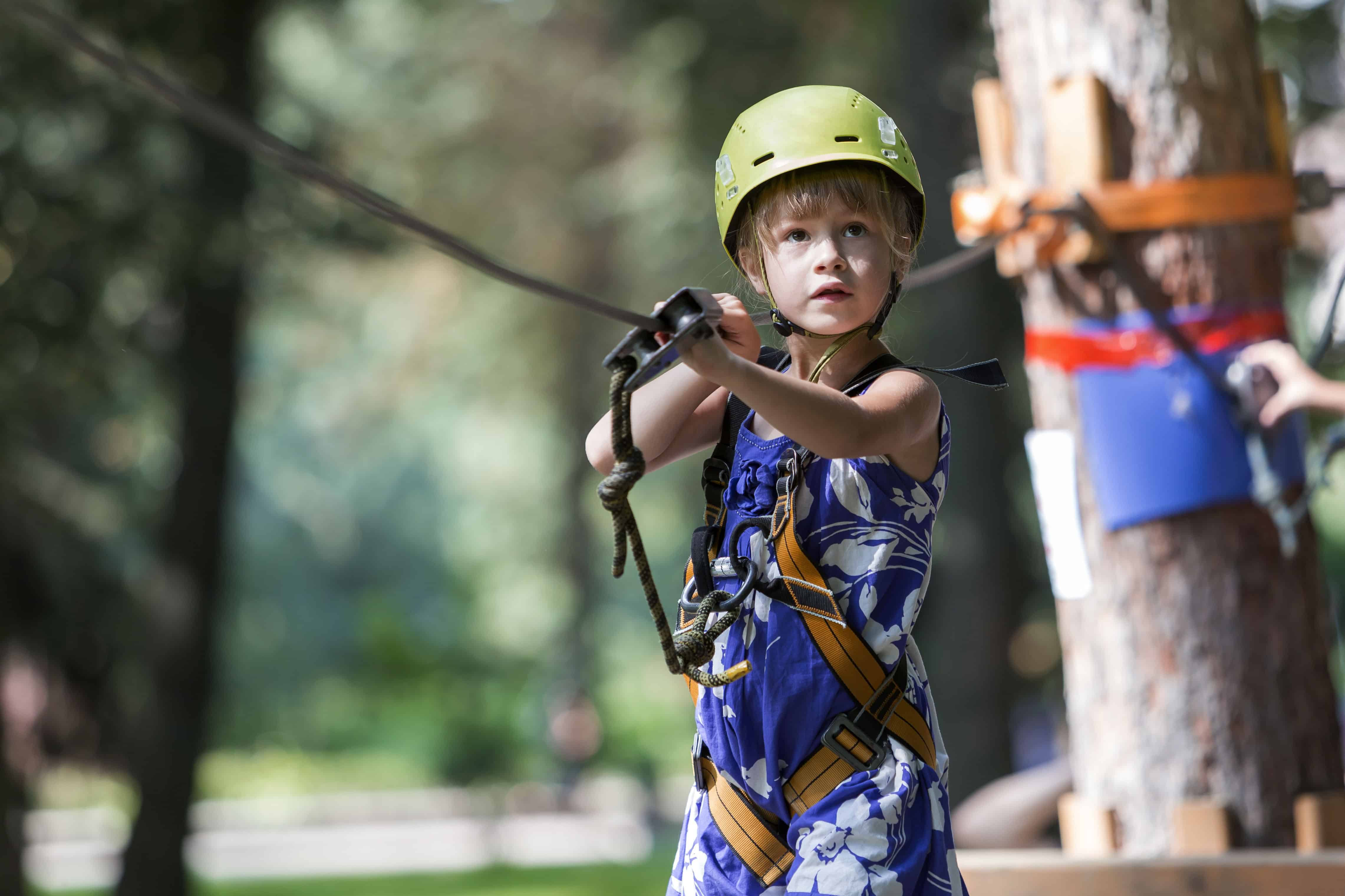 Young child girl in safety harness and helmet attached to cable moves carefully along rope way in recreation park on green bokeh background. Sport, game and leisure concept